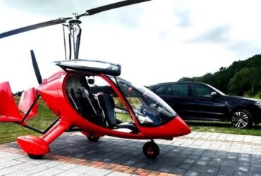 czerwony wiatrakowiec z zabudowaną kabiną pilota stoi na parkingu obok czarnego suva