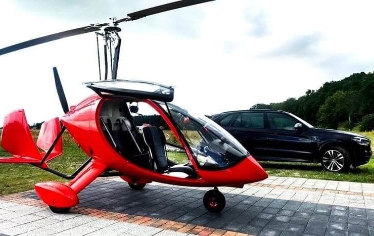 czerwony wiatrakowiec z zabudowaną kabiną pilota stoi na parkingu obok czarnego suva